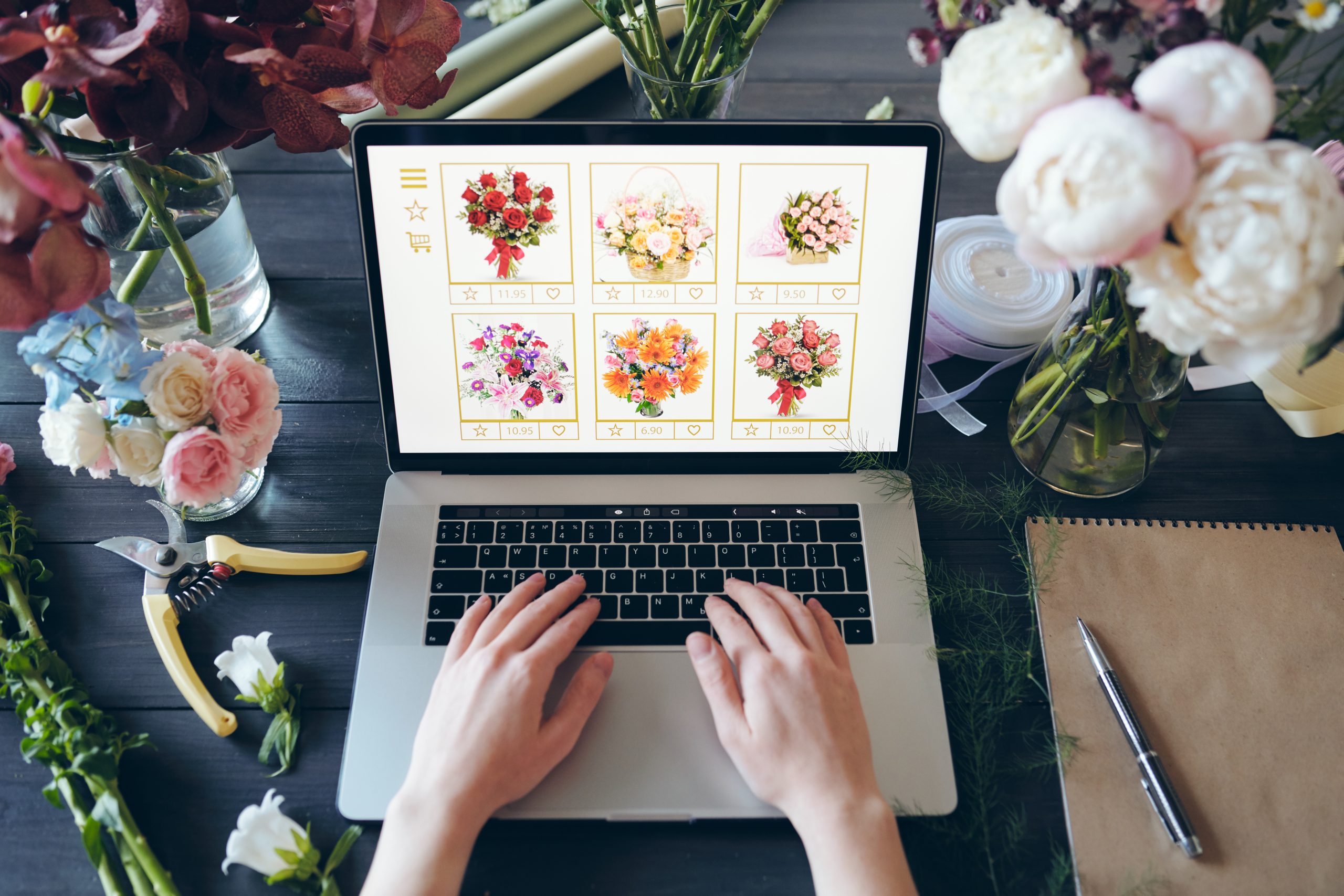 Above view of florist standing at desk with beautiful flowers and hand tools and typing on laptop keyboard