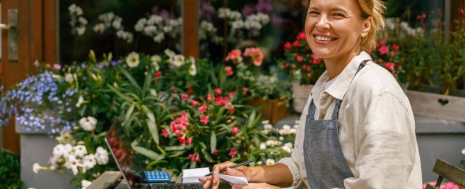 Beautiful flower shop owner wearing apron working on laptop in her store and looking at camera