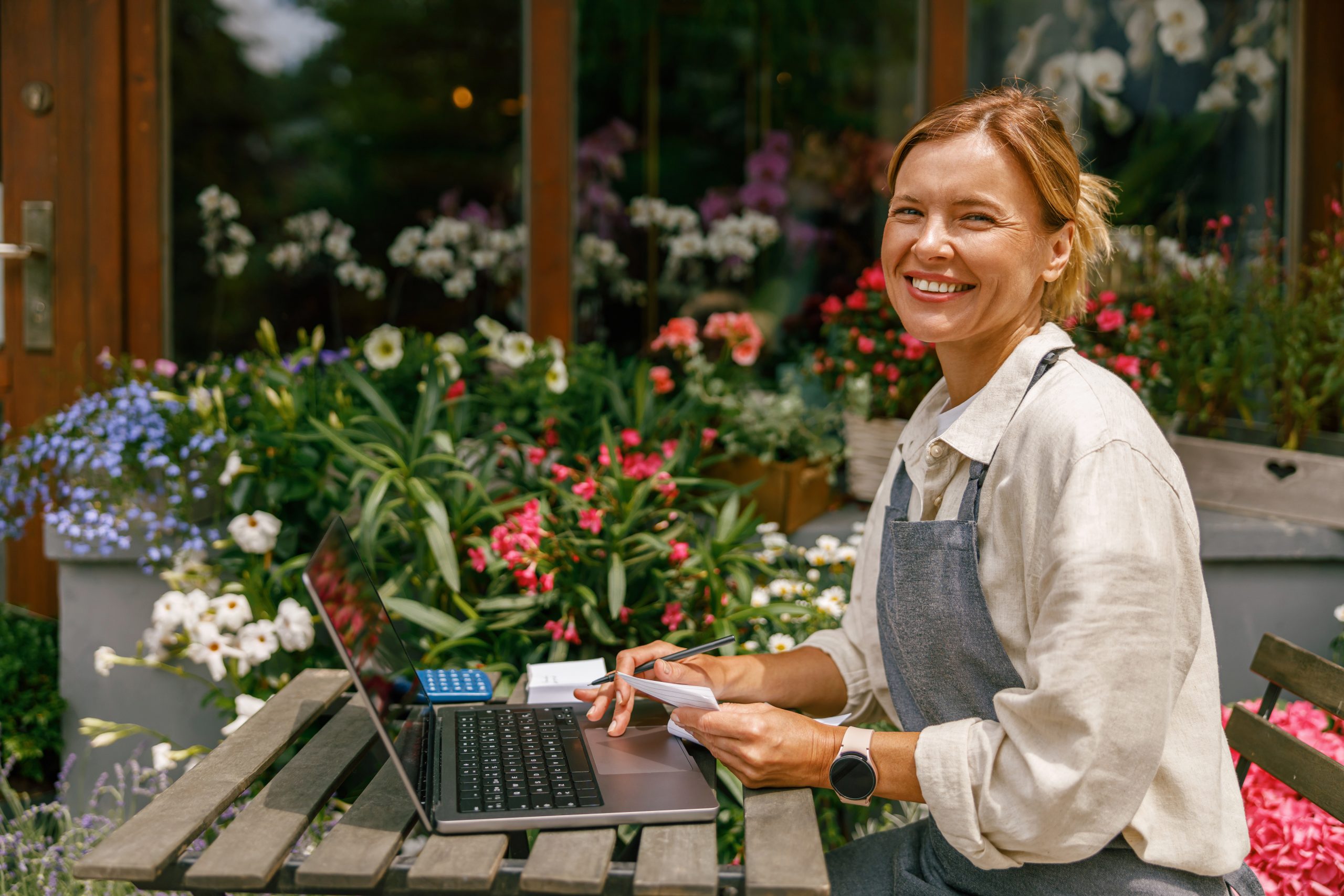 Beautiful flower shop owner wearing apron working on laptop in her store and looking at camera